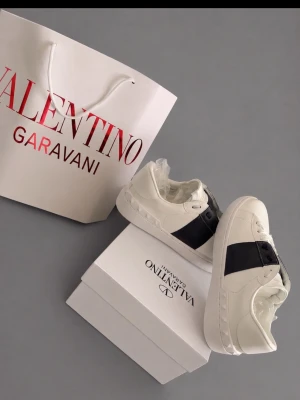 Valentino Garavani vita sneakers - Stilrena vita sneakers från Valentino Garavani med bred svart läderdetalj över sidan. Klassisk låg modell med robust vit sula och diskret logga. Perfekta för dig som vill ha en lyxig och trendig look.