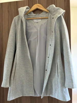 Ljusgrå lång hoodiejacka med huva - Snygg ljusgrå hoodiejacka med huva och långa ärmar. Jackan har en rak passform, knappar framtill och är tillverkad i mjukt material som känns som bomull. Perfekt för lager-på-lager och chill vardagsstil.