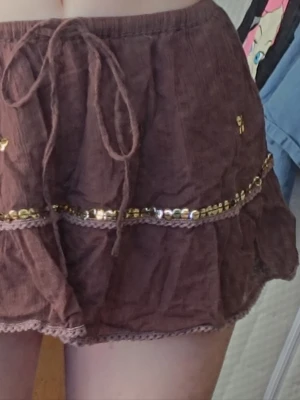 Brun miniskirt med paljettdetaljer - Supersöt brun kortkjol med volanger, snörning i midjan och glittriga paljettdetaljer i guld. Kjolen har en lätt och luftig känsla. Dekorativa sömmar och spetskant nertill ger en extra touch.