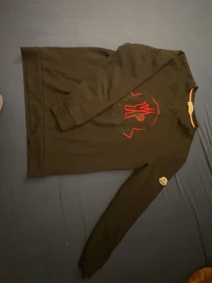 Svart långärmad Moncler t-shirt - Svart långärmad t-shirt från Moncler med stor broderad logga i rött och svart på bröstet samt Moncler-märke på ärmen. Tillverkad i mjuk bomull och har en klassisk passform med rund hals.