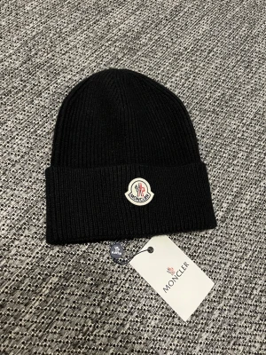 Svart mössa från Moncler - Snygg svart mössa från Moncler med uppvikt kant och klassisk Moncler-logga framtill. Tillverkad i mjukt material som håller dig varm och ger en clean look. Perfekt accessoar för kalla dagar och enkel att matcha med olika outfits.