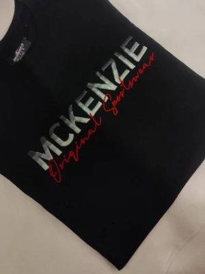 Svart sweatshirt från McKenzie - Svart sweatshirt från McKenzie med stor logga i silver och röd text på bröstet. Klassisk rund halsringning och mjukt material som känns skönt mot huden. Perfekt för en avslappnad och sportig stil.