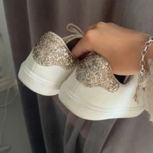 Vita sneakers med glitterdetalj - Säljer ett par vita sneakers från Next med glittriga hälkappor i guld och silver. Skorna har beige snörning och en stjärndetalj på sidan. Tillverkade i syntetmaterial med platt sula och rund tå. 🩷