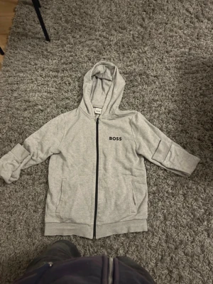 Grå hoodie med dragkedja från BOSS - Snygg ljusgrå hoodie från BOSS med svart logga på bröstet. Hoodien har dragkedja framtill, två fickor och en klassisk huva. Tillverkad i mjukt bomullsmaterial som är skönt att ha på sig. Perfekt för en avslappnad och stilren look.