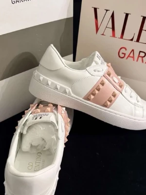Valentino Garavani vita sneakers med nitar - Snygga vita sneakers från Valentino Garavani med ljusrosa detaljer och ikoniska pyramidformade nitar. Skorna har en klassisk låg profil, rund tå och är tillverkade i läder med vita snören. Perfekta för dig som vill sticka ut med en lyxig touch.