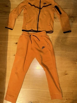 Orange mjukisbyxor från Nike - Snygga orange mjukisbyxor från Nike med svart logga på låret och dragsko i midjan. Byxorna har ribbade muddar vid bensluten och praktiska fickor. Perfekta för chill eller träning, tillverkade i mjukt material för maximal komfort. Nike tech som har blivit försmå. Köpte byxorna och huddien tillsammans för 3000kr (ÄKTA) 