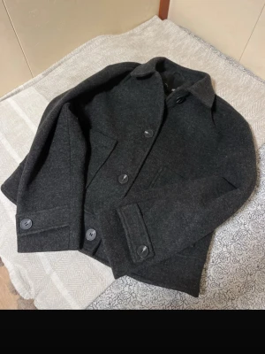 Mörkgrå kappa - Stilren mörkgrå kappa. Jackan har två stora fickor och raka ärmar med knappdetaljer vid ärmsluten. Nyskick! 