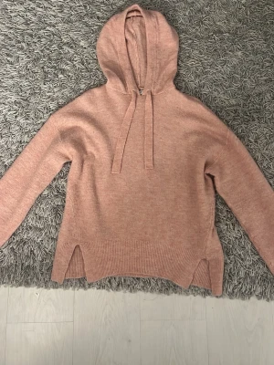 Stickad rosa stocklmstil hoodie med slitsar och huva - Mysig stickad hoodie i ljusrosa med huva och dragsko. Tröjan har långa ärmar och slitsar på sidorna vid nederkanten. Perfekt för en avslappnad och trendig look.