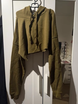 Olivgrön cropped hoodie - Säljer en olivgrön cropped hoodie med huva och långa ärmar. Hoodien har en avslappnad passform, dragsko i huvan och är gjord i mjukt sweatshirtmaterial. Perfekt för en chill och trendig look.