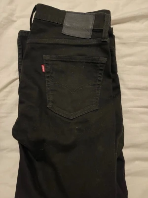 Svarta Levi's jeans med rak passform - Säljer ett par svarta jeans från Levi's med klassisk femficksdesign och rak passform. Jeansen har den ikoniska röda Levi's-lappen på bakfickan och ett svart läderpatch i midjan. Perfekta till en avslappnad streetwear-look.