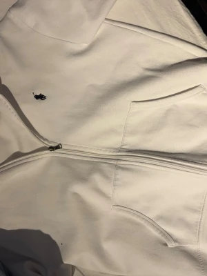 Vit hoodie med dragkedja från Polo Ralph Lauren - Snygg vit hoodie med dragkedja från Polo Ralph Lauren. Klassisk design med huva, två fickor framtill och den ikoniska broderade loggan på bröstet. Perfekt för en clean och stilren look. Mjuk bomullskvalitet som känns skön mot huden.