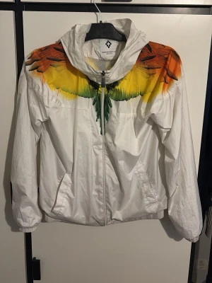 Marcelo Burlon Vindjacka - Säljer en vit Marcelo burlon vindjacka med dragkedja och huva. Jackan har ett unikt tryck av färgglada fjädrar i orange, gul och grön över axlar och bröst, som fortsätter på ryggen. Två fickor framtill och muddar. Perfekt för dig som vill sticka ut.