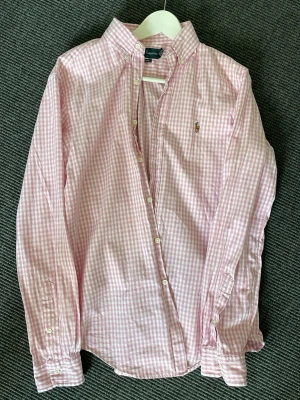 Rosa rutig skjorta från Ralph Lauren - Klassisk rosa och vit rutig skjorta från Ralph Lauren med button-down krage och broderad logga på bröstet. Skjortan har långa ärmar och knäppning framtill. Perfekt för en stilren och avslappnad look.