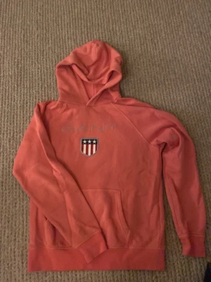 Rosa hoodie från GANT med logga - Snygg rosa hoodie från GANT med broderad logga och ett sköldmärke framtill. Klassisk huva och stor magficka. Perfekt för en avslappnad och trendig stil. Mjuk bomullskänsla och långa ärmar.