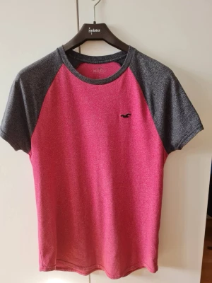 Röd och svart t-shirt från Hollister strl S - Snygg t-shirt från Hollister i en fräsch röd färg med kontrasterande svarta ärmar. Den har en liten logga på bröstet och korta ärmar. Storlek S. Kan hämtas i Vallentuna.