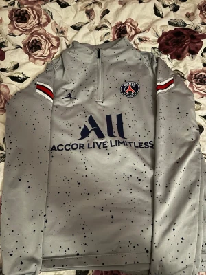 Grå PSG träningströja Jordan - Snygg grå träningströja från PSG och Jordan med halv dragkedja, mörkblå logga och text, samt svarta prickar över hela tröjan. Röda, vita och blå detaljer på ärmarna. Perfekt för fotbollsträning eller chill. Materialet är mjukt och stretchigt, troligen polyester.