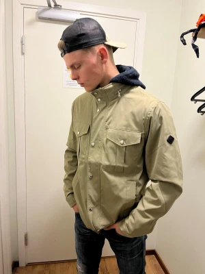 J.LINDBERG  jacka - Snygg beige jacka med två bröstfickor med lock och tryckknappar. Klassisk passform och diskret logotyp på ärmen. Materialet är slitstarkt och passar perfekt till lager-på-lager. Perfekt för dig som gillar stilren och funktionell design.