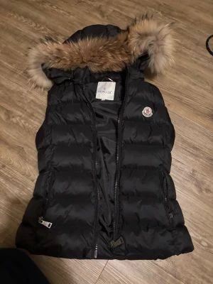 Svart dunväst med fuskpäls från Moncler - Snygg svart dunväst från Moncler med fluffig huva kantad av fuskpäls. Västen har två dragkedjefickor, Moncler-logga på bröstet och quiltad design. Perfekt för lager på lager och ger en cool streetstyle-vibe.