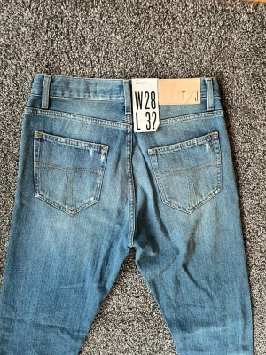 Blå jeans från Tiger of Sweden - Säljer ett par klassiska blå jeans från Tiger of sweden storlek W28 L32. Jeansen har raka ben, två bakfickor och snygga slitningar för en avslappnad look. Tillverkade i mjukt denimtyg med ljus tvätt och kontrastsömmar. Perfekta för en casual stil.