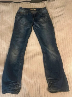 Blå bootcut jeans - Säljer ett par blå bootcut jeans från G-Unit med slitningar och ljusa partier på låren och knäna. Jeansen har klassisk femficksdesign, låg midja och detaljerade slitningar. Har bytt knapp till en avtagbar( kan skicka med extra sånna ) passar på mig som är 169