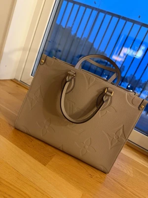 Beige Louis Vuitton handväska - Snygg beige handväska från Louis Vuitton med klassiskt monogrammönster i präglat läder. Väskan har både korta handtag och långa axelremmar i samma färg, samt guldfärgade metalldetaljer. Perfekt för dig som vill ha en stilren och lyxig accessoar.