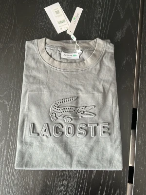 Grå Lacoste t-shirt med logga - Snygg grå t-shirt från Lacoste med stor upphöjd krokodillogga och text på bröstet. Klassisk rund halsringning och korta ärmar. Tillverkad i mjuk bomull för en skön känsla. Perfekt för en clean och stilren look.