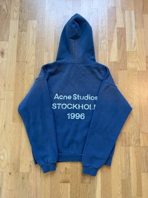 Blå hoodie Acne Studios Stockholm - Blå hoodie från Acne Studios med stor huva och tryck på ryggen där det står 'Acne Studios STOCKHOLM 1996' i vitt. Klassisk passform med ribbade muddar vid ärmslut och midja. Perfekt för dig som gillar streetwear och vill ha något stilrent men ändå unikt.