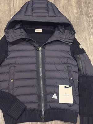 Mörkblå pufferjacka från Moncler - Snygg mörkblå cardigan från Moncler med huva och stickade ärmar. Jackan har två framfickor med tryckknappar, dragkedja framtill och ribbade muddar. Klassisk Moncler-logga på ärmen och quiltad design ger en modern look. Perfekt för kalla dagar.