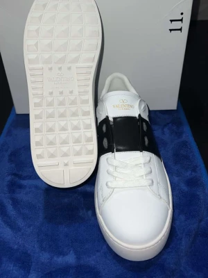 Valentino Garavani vita sneakers - Stilrena vita sneakers från Valentino Garavani med svart läderdetalj och nitar på sidorna. Klassisk låg modell med vita skosnören och diskret logga på plösen. Yttermaterial i slätt läder och gummisula med rutmönster undertill.