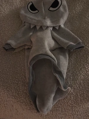 Grå hajdräkt för husdjur - Rolig grå hajdräkt för husdjur med huva som har stora ögon och vita hajtänder. Dräkten har fyra ben och mörkare grå kantband. Perfekt för att klä ut din hund eller katt till haj. Ny utan prislapp! Använd en gång inomhus! Kontakta gärna om ni har frågor om storlek eller annat!!