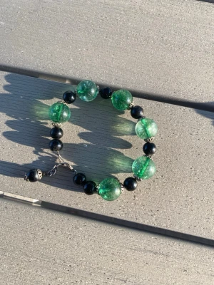 Armband med gröna och svarta pärlor - Snyggt egentillverkat armband med stora gröna glaspärlor och mindre svarta pärlor. Armbandet har en metallkedja och dekorativa detaljer mellan pärlorna. Längd på armband ca 20 cm. 