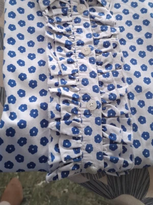 Vit blommig skjorta med volang - Vit skjorta med blått blommönster och volangdetaljer längs knappraden. Långärmad och tillverkad i mjukt bomullstyg. Perfekt för dig som gillar färg och mönster! Bystmått: från armhåla till armhåla 48 cm t: från sida till sida längst ner 53 cm Längd: 60 cm