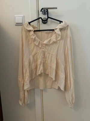 Krämvit volangblus med spetsdetaljer - Romantisk krämvit blus med volangkrage och knappar framtill. Blusen har långa ärmar och fina spetsdetaljer längs bröstet och ärmarna. Tillverkad i ett tunt, lätt tyg som ger en luftig känsla. Perfekt för dig som gillar en feminin och vintageinspirerad stil.