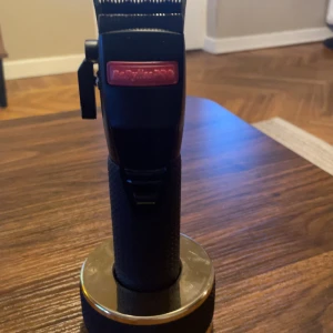 BabylissPRO hårtrimmer svart - Elektrisk hårtrimmer från BabylissPRO i svart med texten 'DLC FADE' på skärhuvudet. Trimmern har ett greppvänligt handtag och står på en rund laddstation i metall. Perfekt för att fixa snygga fades och detaljerade frisyrer.