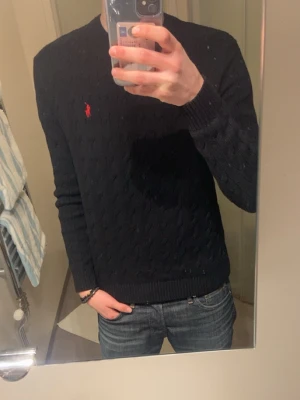 Mörkblå kabelstickad tröja Polo Ralph Lauren - Säljer en mörkblå kabelstickad tröja från Polo Ralph Lauren med den klassiska röda loggan broderad på bröstet. Tröjan har rund halsringning, ribbade muddar och är tillverkad i mjukt material. Perfekt för dig som vill ha en stilren och tidlös look.