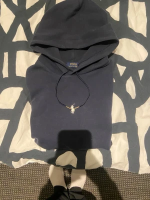 Mörkblå hoodie från Polo Ralph Lauren - Säljer en mörkblå hoodie från Polo Ralph Lauren med klassisk huva och dragsko. Ikoniska Polo-loggan broderad på bröstet. Hoodien är i mjukt bomullsmaterial och har en avslappnad passform, perfekt för en chill och stilren look.litet hål i magen därmed priset .