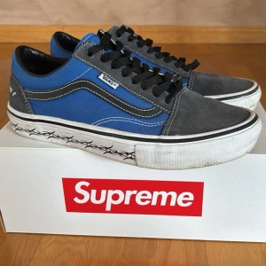 Vans x Supreme blå/grå sneakers - Säljer ett par Vans x Supreme sneakers i blått och grått med svarta detaljer. Skorna har klassisk låg siluett, svart-vit sula med taggad design och Supreme-logga bak. Ovandelen är i mocka och canvas, med svarta skosnören och vita sömmar.