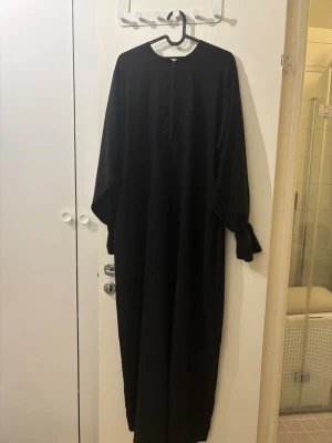 Svart elegant Sara abaya från Tasnim collections - Superfin svart abaya från tasnim collections. Använt fåtal gånger, nyskick med dragkedja och fickor😍  Modell: Sara abaya Färg: svart W2 Längd: 140cm  Har även en annan klänning på min sida från Tasnim collections som är i lika bra skick, kika gärna🥰