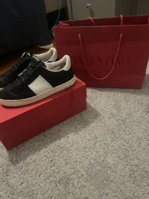 Valentino flycrew  svartvita sneakers - Säljer ett par snygga svartvita sneakers från Valentino. Skorna har en låg silhuett, vita detaljer på sidan och små silverfärgade nitar på sulan. Tillverkade i mocka med klassisk snörning och vit yttersula. Perfekta för dig som gillar exklusiv streetstyle.
