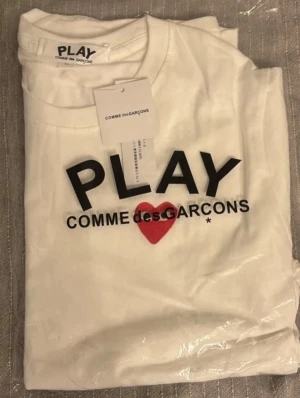Vit t-shirt Comme des Garçons Play - Vit t-shirt från Comme des Garçons Play med svart tryck och rött hjärta på bröstet. Klassisk rund halsringning och korta ärmar. Tillverkad i mjuk bomull och har en stilren, modern look med tydlig logga framtill.