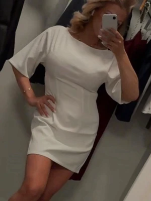 Vit kortklänning med vida ärmar - En stilren vit kortklänning med rund halsringning och vida, korta ärmar. Klänningen har en enkel och rak passform som ger ett modernt uttryck. Perfekt för dig som gillar minimalistisk design och vill ha något lätt och bekvämt.