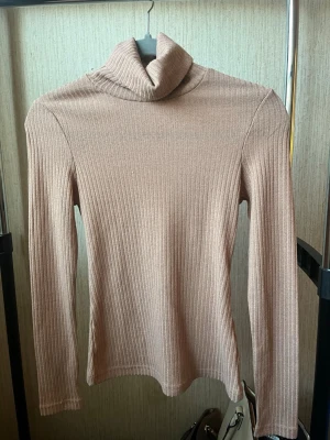 Beige ribbad polotröja - Säljer en beige ribbad polotröja med lång ärm och hög krage. Tröjan har en smal passform och är tillverkad i ett mjukt, stretchigt material som sitter snyggt på kroppen. Perfekt för lager-på-lager och enkel att matcha med olika outfits.
