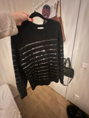 Svart stickad tröja med paljettränder - Säljer en svart stickad tröja med glittriga paljettränder över hela tröjan. Tröjan har rund halsringning, långa ärmar och ribbade muddar vid ärmslut och nederkant. Perfekt för dig som vill ha något extra till din outfit.