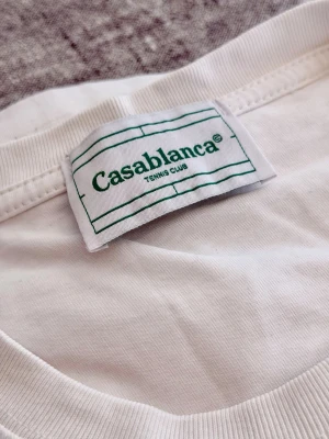 Vit t-shirt från Casablanca - Vit t-shirt från Casablanca med rund halsringning ochi blått tryck på bröst/rygg. T-shirten har en clean look och är tillverkad i mjuk bomull. Perfekt för dig som gillar stilrena plagg med en twist. Inköpt NK hösten 2025, nypris: 1899:-