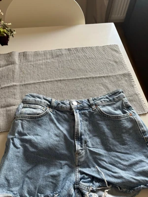 Ljusblå jeansshorts midwaist S - Säljer ett par ljusblå jeansshorts i midwaist-modell. Klassisk femficksdesign med råa, fransiga benslut och knappgylf. Perfekta till sommaren och lätta att matcha med olika toppar.