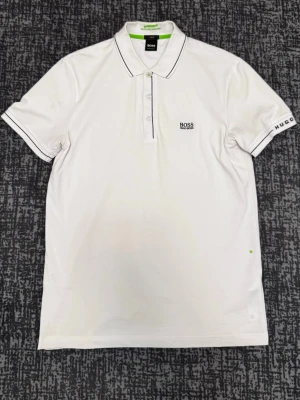 Vit pikétröja från Hugo Boss - Vit pikétröja från Hugo Boss med klassisk krage, knäppning framtill och diskreta svarta detaljer på krage och ärmslut. Broderad logga på bröstet och märkesnamn på ärmen. Perfekt för en stilren look.