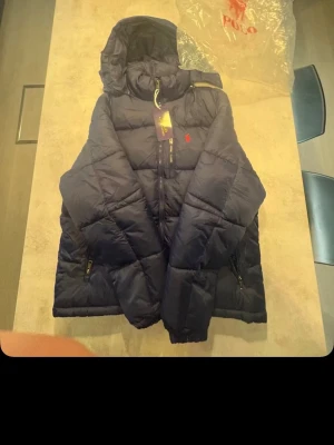 Mörkblå dunjacka från Polo Ralph Lauren - Snygg mörkblå dunjacka från Polo Ralph Lauren med huva, dragkedja och två fickor med dragkedja. Jackan har justerbar dragsko i nederkant och ikonisk röd logga på bröstet. Perfekt för kalla vinterdagar och riktigt skön att bära. Den är lite trasig där man kan spänna den