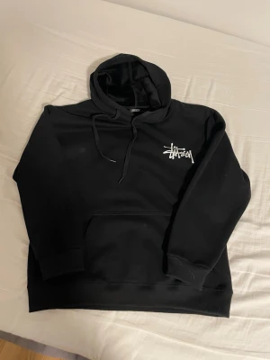 Svart Stüssy hoodie - Svart hoodie från Stüssy med stor vit logga på ryggen och mindre logga på bröstet. Perfekt streetwear-plagg i mjukt material, riktigt skön på insidan. Sparsamt använd, inga defekter. Nypris ca 1900kr. Storlek L men passar bättre till M! Hör av er vid frågor till exempel om mått!