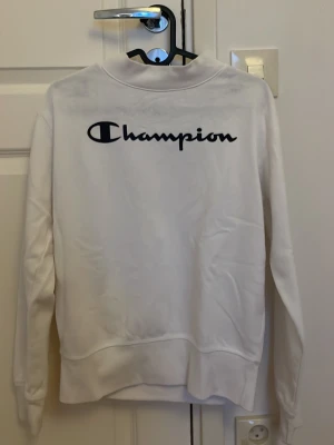 Vit sweatshirt från Champion - Vit sweatshirt från Champion med svart logotyp tryckt på ryggen. Klassisk modell med rund halsringning och långa ärmar. Tillverkad i mjukt material som passar perfekt för en avslappnad stil. Inte använd alls, den är i barn storlek