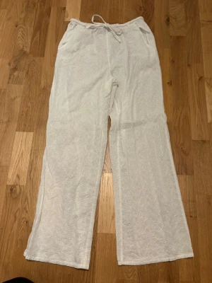 Vita broderade linnebyxor med dragsko - Snygga vita linnebyxor med broderade små blommor och avslappnad passform. Byxorna har dragsko i midjan och raka ben, perfekta för en chill och luftig stil. Materialet är tunt och andas, vilket gör dem grymt sköna under varma dagar.